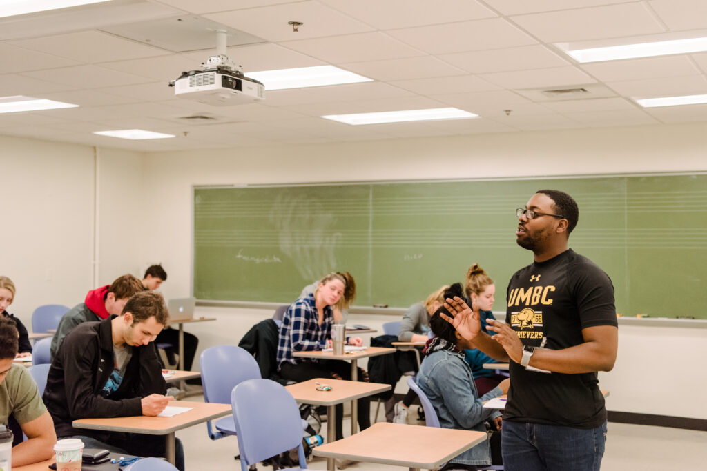 Student in UMBC t-shirt speaks in front of a class, while other students take notes.