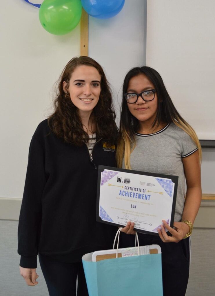 Two young women pose for a portrait, one holding a gift back and achievement certificate.