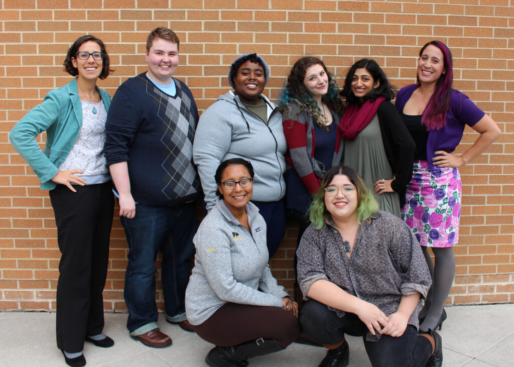 Eight people in their 20s and 30s, of diverse identities, stand in two rows in front of a brick wall.