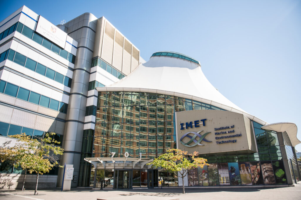 A view of a building with a tall glass entrance. The building has the letters IMET written in navy blue with an outlines of two purple and green fish underneath.