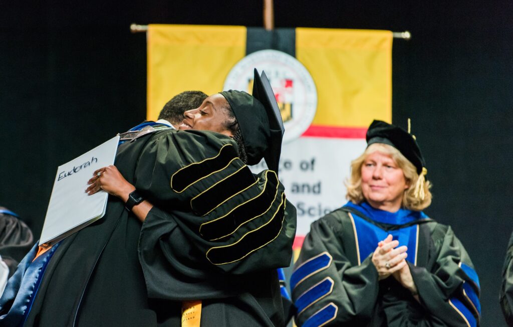 Man in highly decorated commencement robe hugs student speaker. Behind them, another person in highly decorated robe claps.