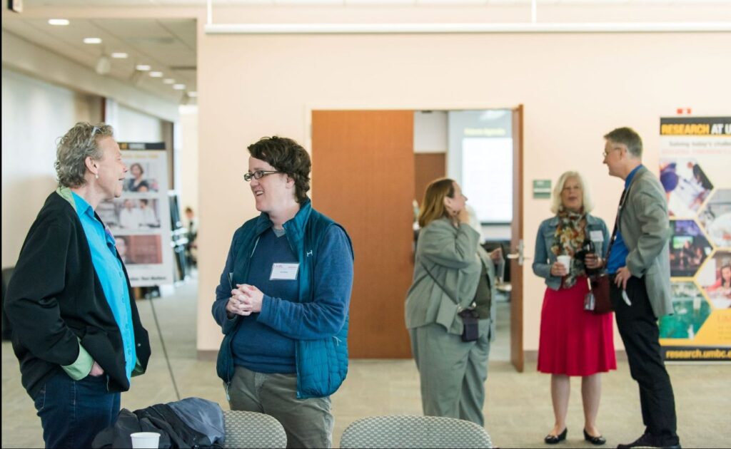 Two faculty members talking at a university research event, with three additional faculty members having a separate conversation in the background.