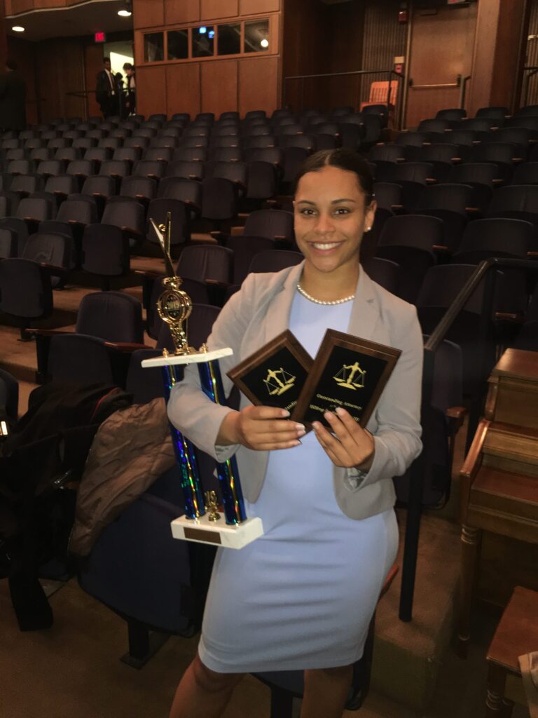 Young woman with dark hair wearing a grey blazer and a light blue dress smiles at camera while holding two plaques and one trophy while standing in an auditorium.