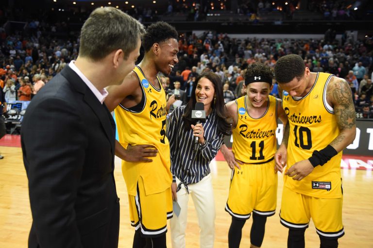 A reporter standing on court with three UMBC basketball players.