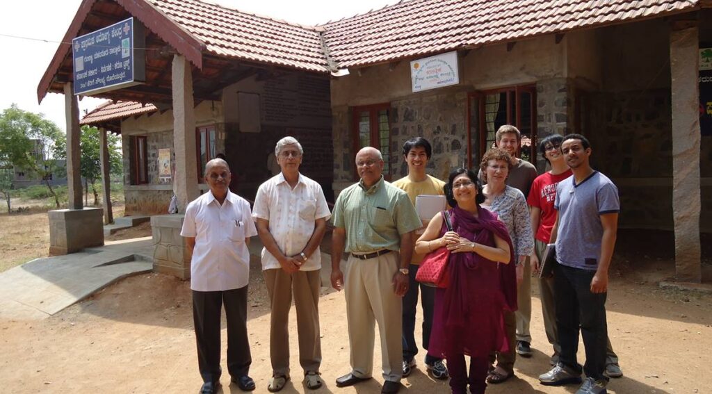 A group of nine people of different ages stand in a group in front of a one floor stone house with terracotta