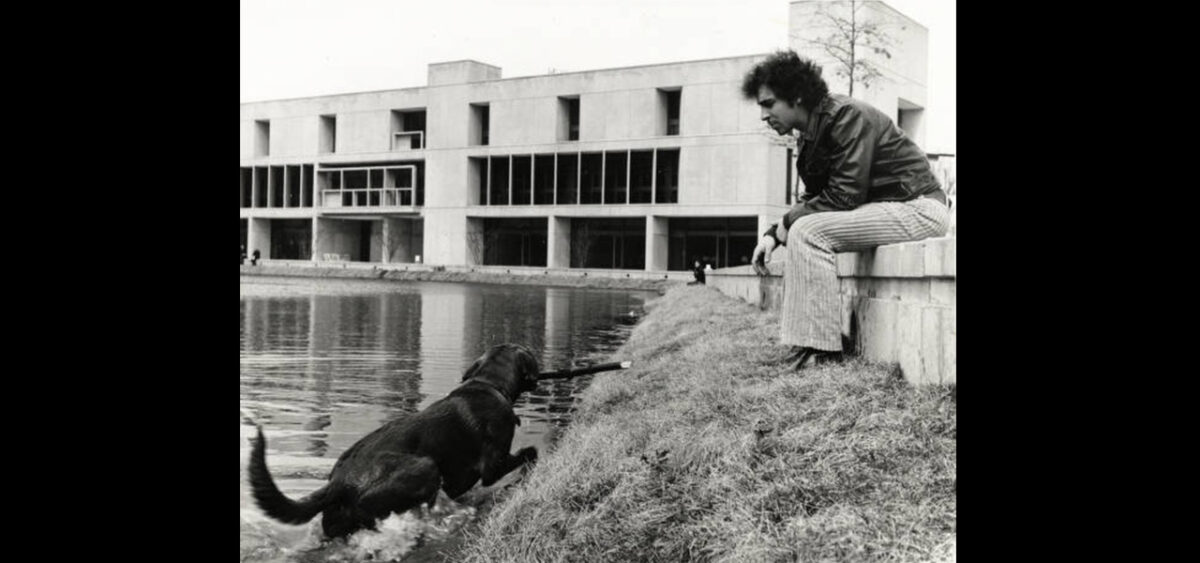 in a black and white photo a man sits on a low wall while a dog comes out of a pond with a stick in its mouth