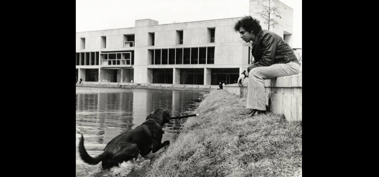 in a black and white photo a man sits on a low wall while a dog comes out of a pond with a stick in its mouth