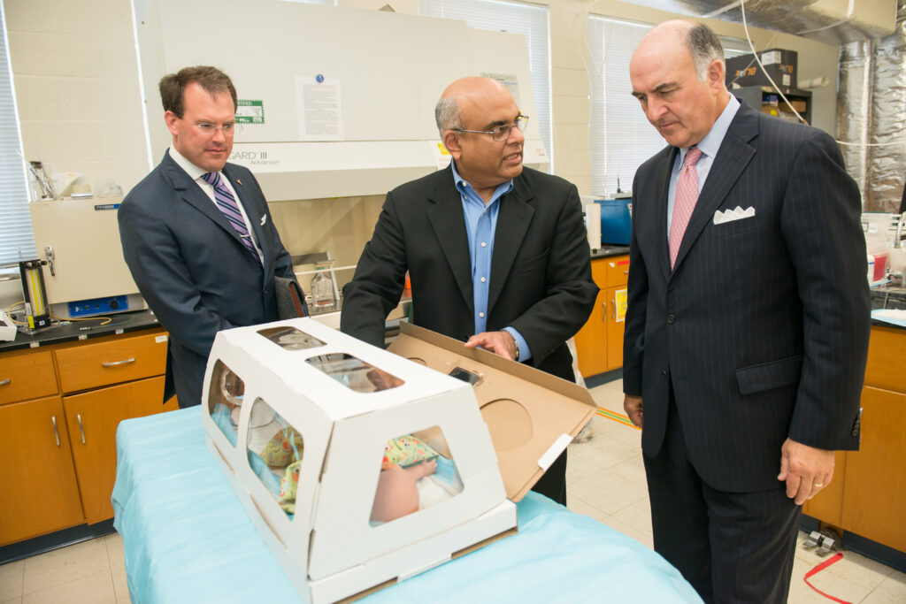 Three men in suits are in a lab gathering around a table looking at a cardboard box with a baby doll inside simulating an incubator.