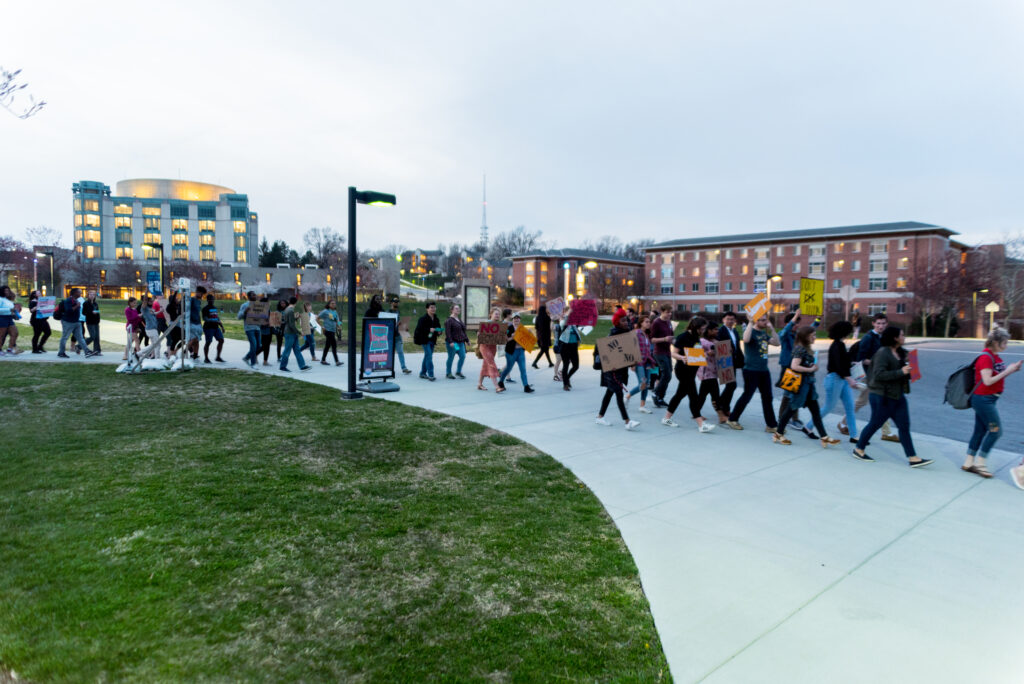 A group of students and other university community members walks through the university, carrying signs in support of women and survivors of sexual violence.