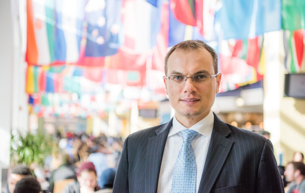 Man in suit and tie stands in front of  several international flags