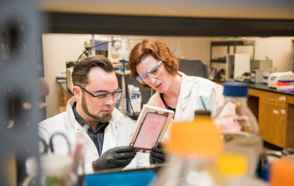 White man with dark beard and white woman with red hair look at a sample in a lab. Both wear lab coats and goggles.