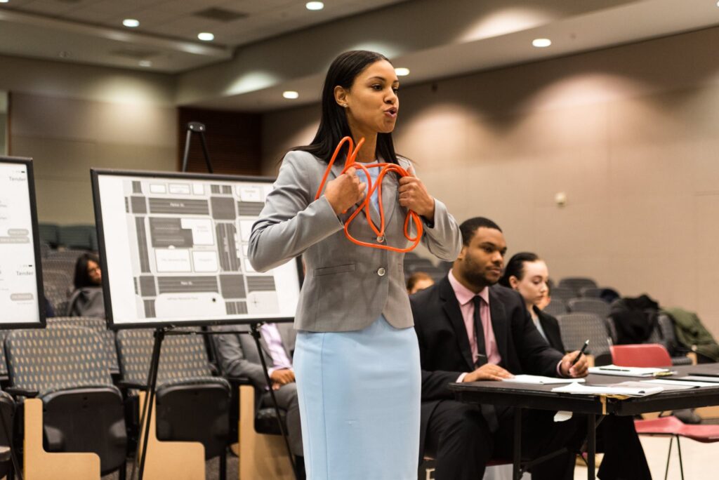 Young woman stands wearing a blue dress and gray blazer, delivering remarks while holding a prop