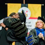 University president hugs undergraduate commencement speaker in congratulations following her remarks, while colleagues clap, all in graduation attire.