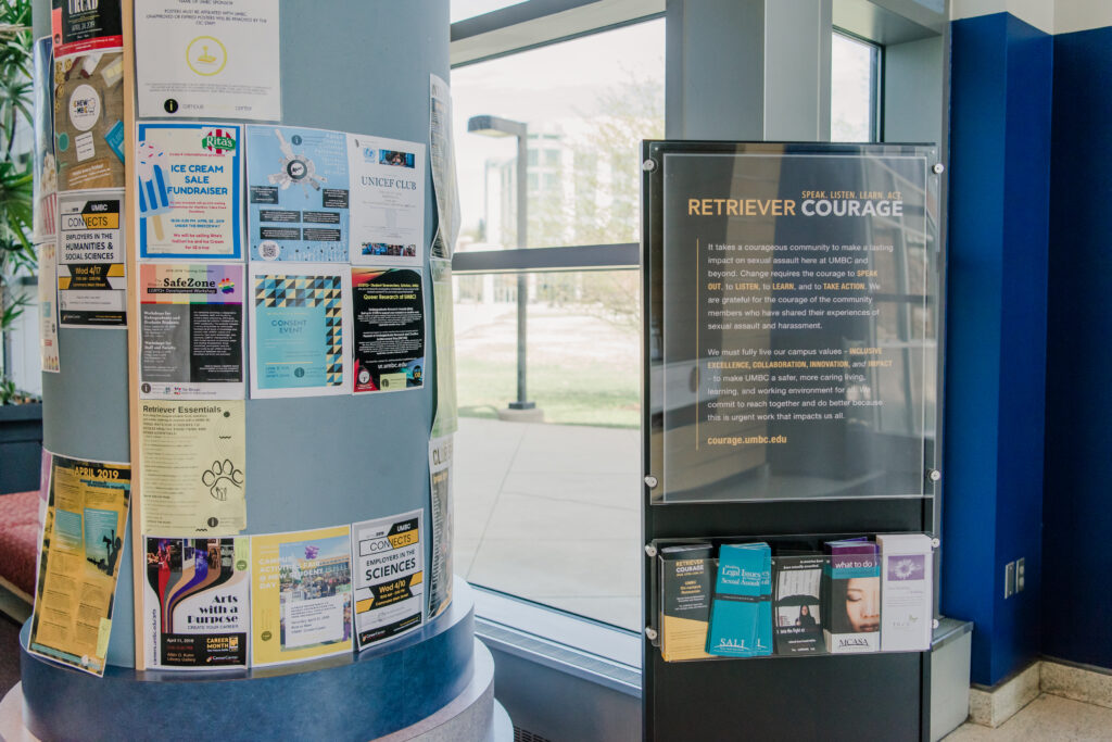 A display board in a university building shares information on Retriever Courage, with space for informational pamphlets below. Toward its left is a pillar with flyers promoting additional events and resources.