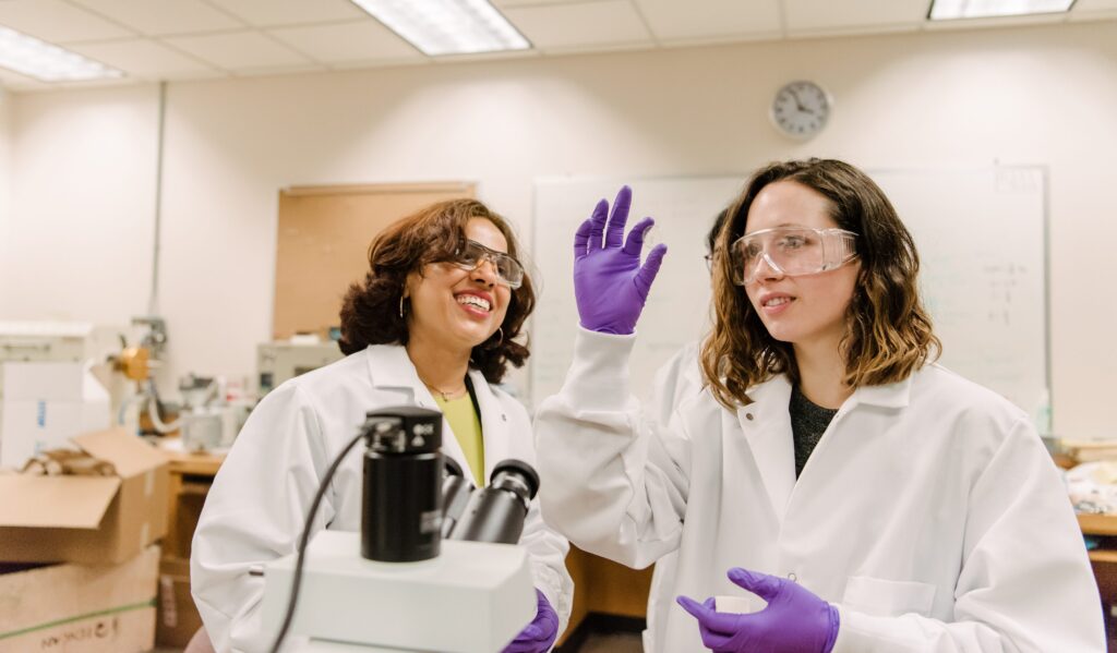Two women work in a lab, with the student looking at a slide while her professor looks on; they wear gloves and goggles