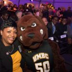 Maryland House Speaker Adrienne Jones, in gold and black, poses with UMBC Retriever mascot, in front of a crowd of people, during an evening celebration.