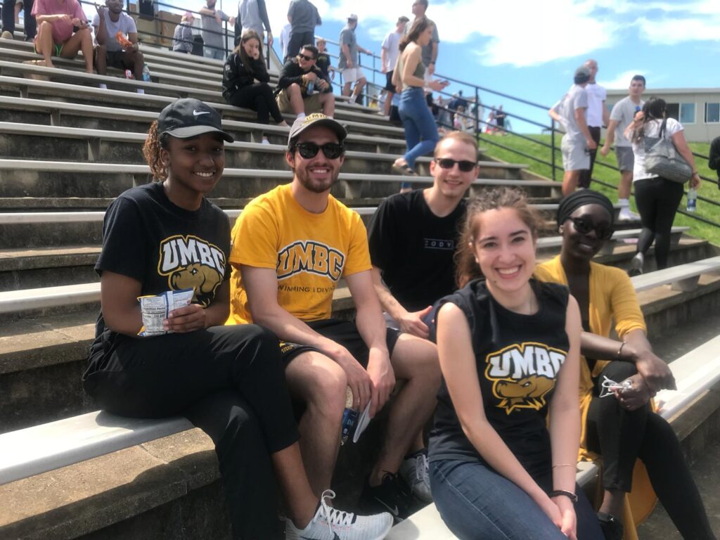 Five students wearing UMBC t-shirts sit, smiling, in the stands at an outdoor athletic game.