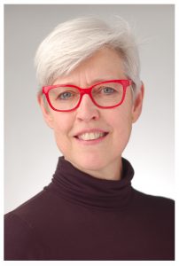 Portrait of woman with short white hair and bright red glasses, in a turtleneck shirt.