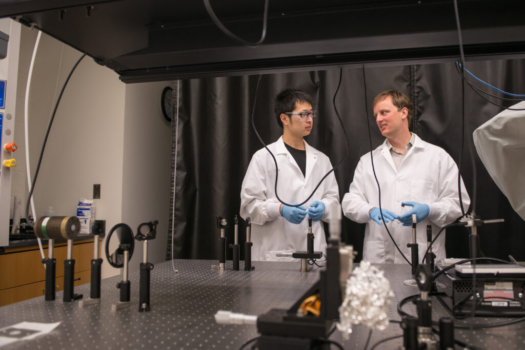 Two scientists in white coats, glasses, and gloves stand in a physics lab.