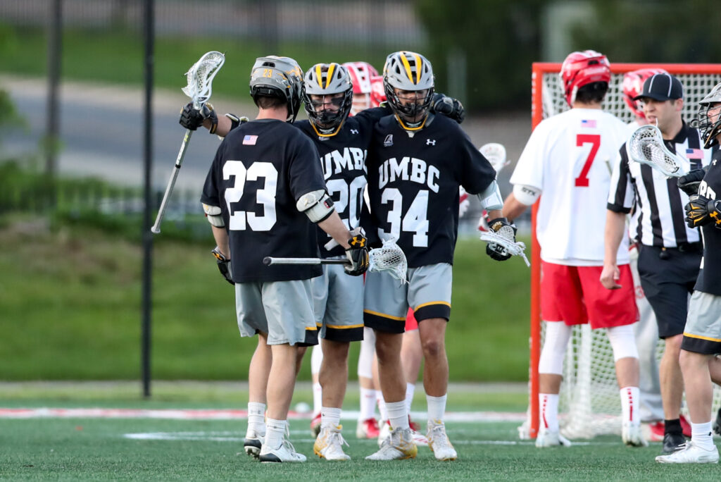 A group of UMBC men's lacrosse players in black, gold, and gray uniforms embraces each other on a field.