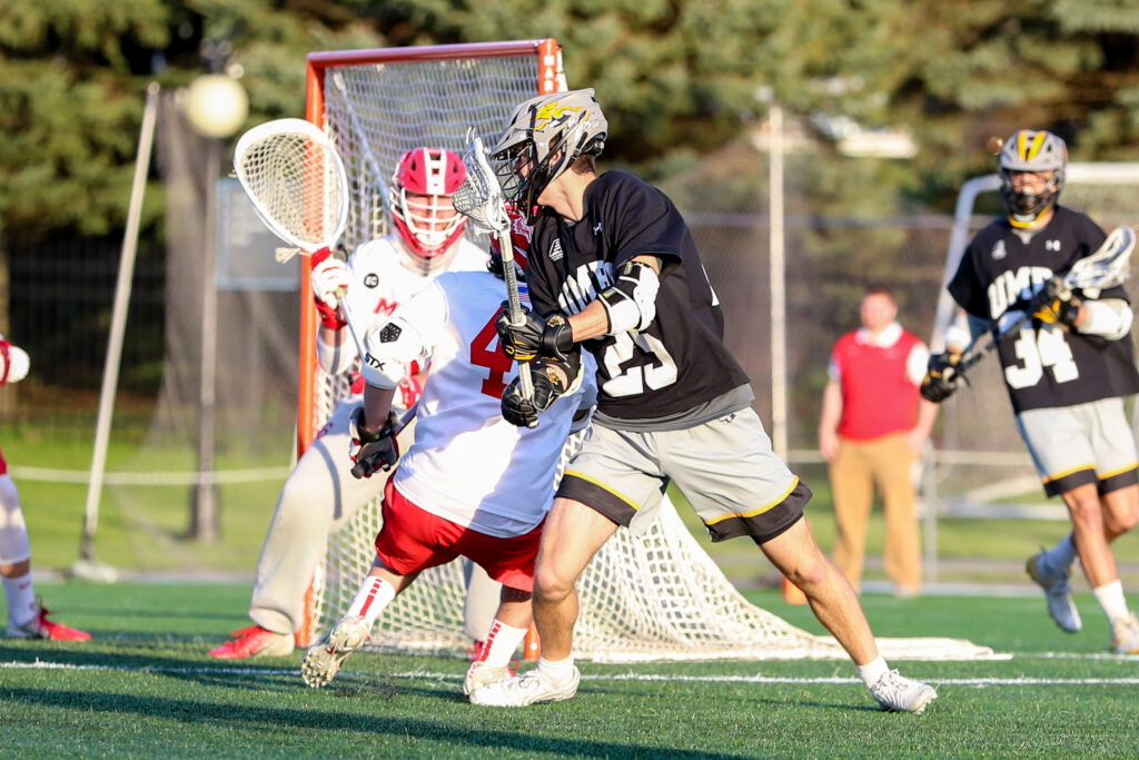 UMBC lacrosse player in black, gold and gray uniform and Marist lacrosse player in red and white uniform, on the field.