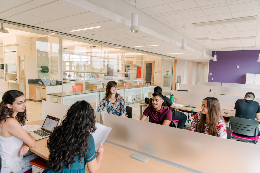 Fives students sit at tables in a shared meeting and work space in a brightly lit new building.