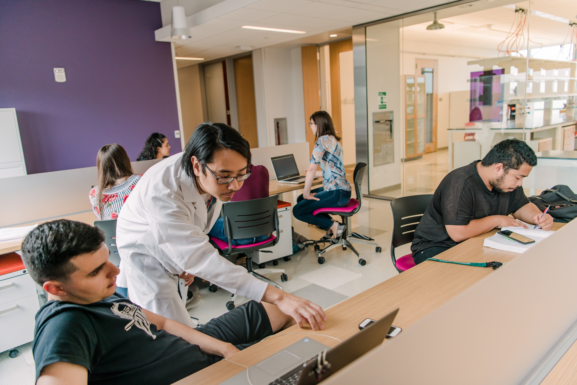 People work at desks in ILSB with student in lab coat helping out