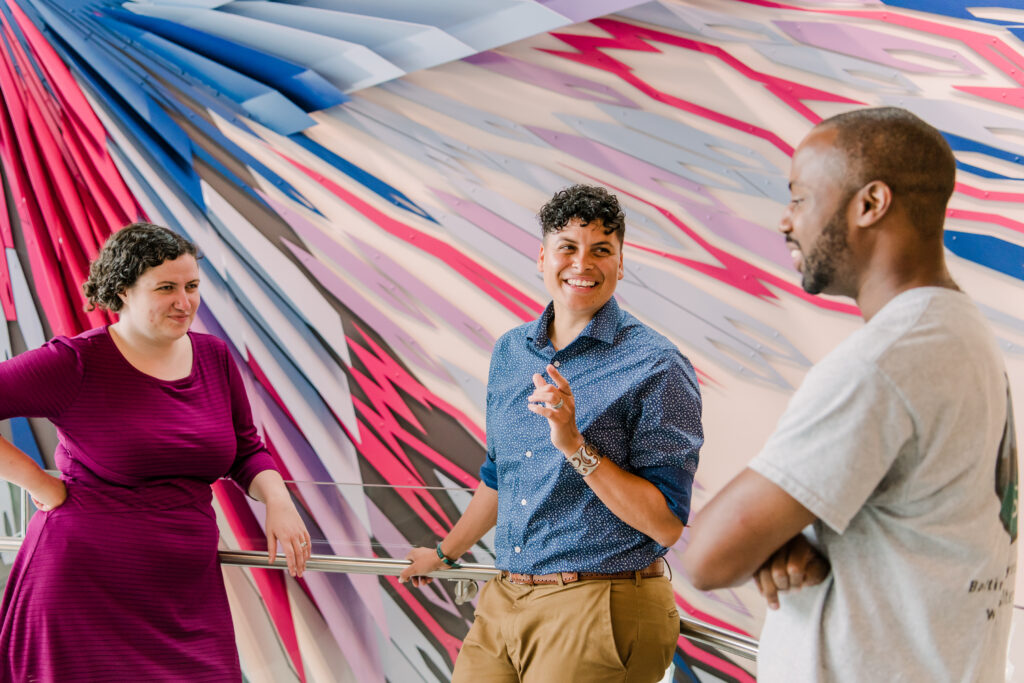 Three people in conversation below a colorful wall art installation