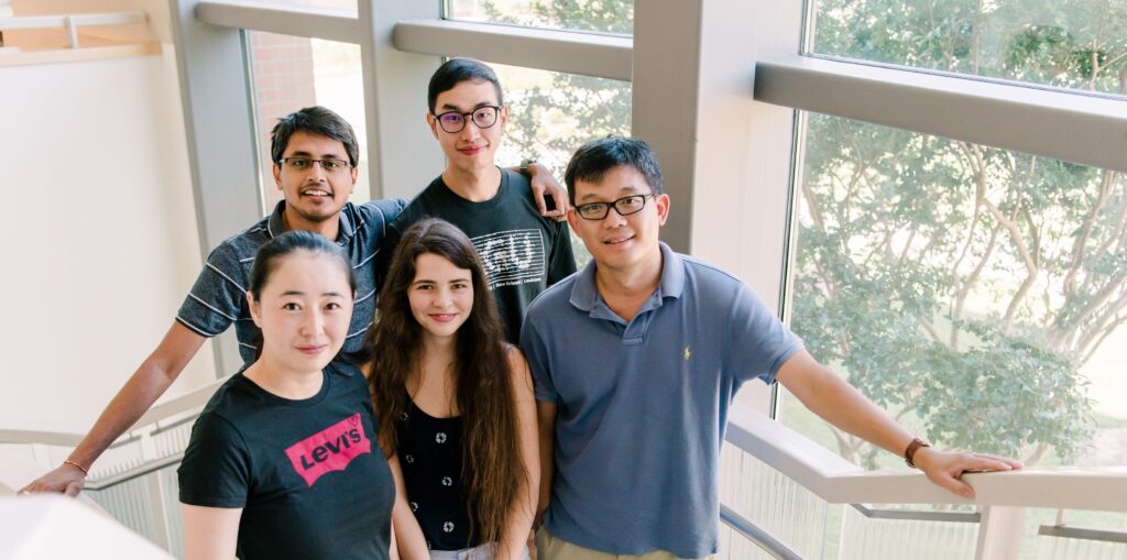 Group of five people stands in front of a window. They are smiling.