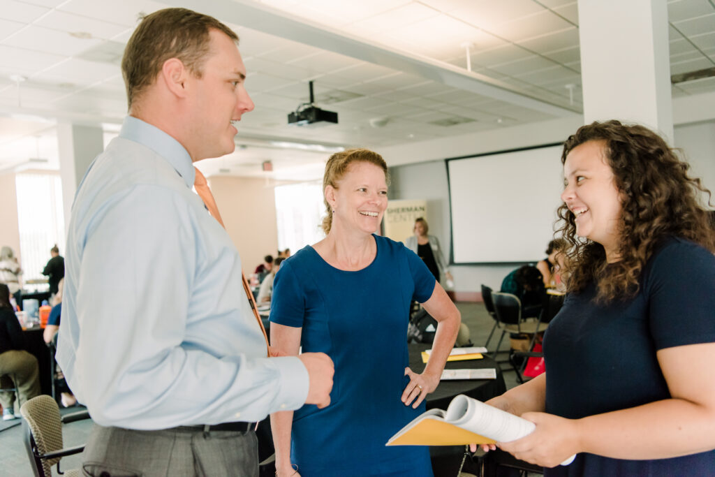 Three people stand, talking to one another, inside a meeting space, in office attire.