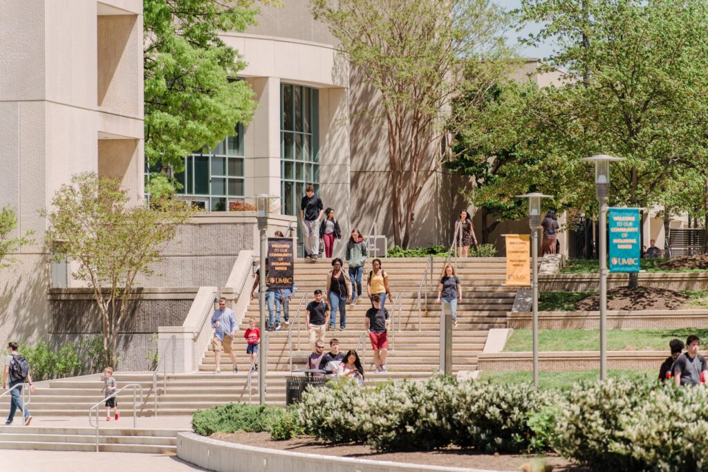 Students walk down stairs in front of a library, surrounded by spring plants, in the sunshine.