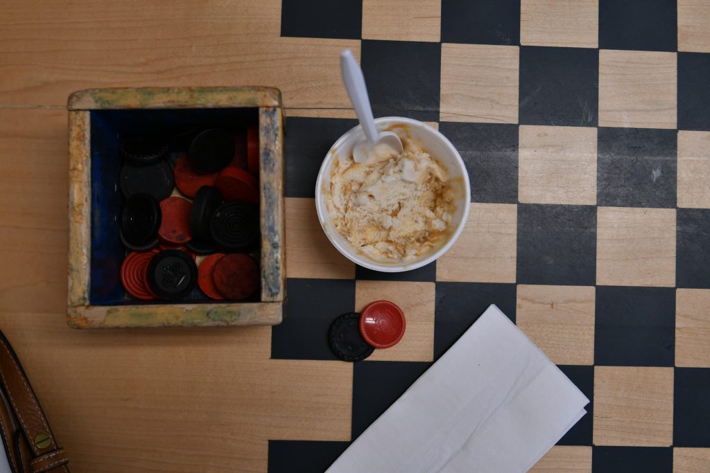 A cup of vanilla butterscotch ice cream atop a wooden checkerboard table. Beside it is a wooden box of black and red checkers pieces.