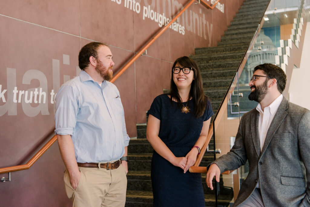 A woman wearing a navy blue dress stands in between two men wearing dress shirts, pants, and jacket while standing at the bottom of the staircase with a brown wall with words behind them.