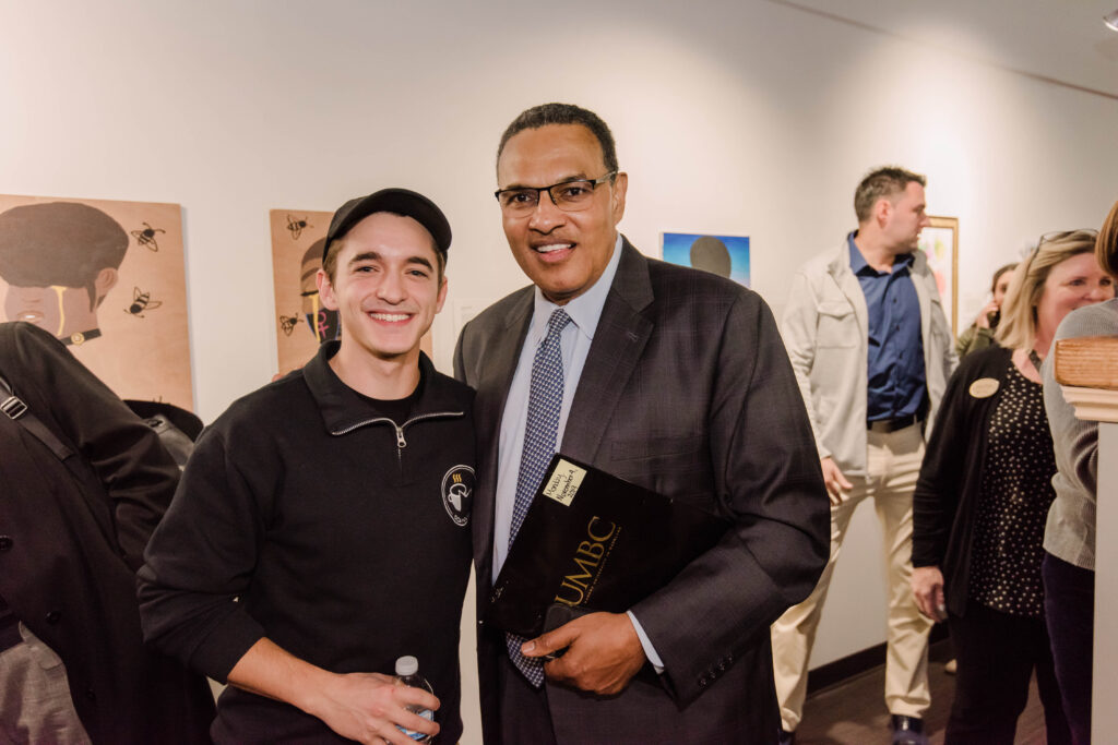 Student in OCA Mocha-branded sweater and cap poses with university president in suit and tie, in front of artwork/