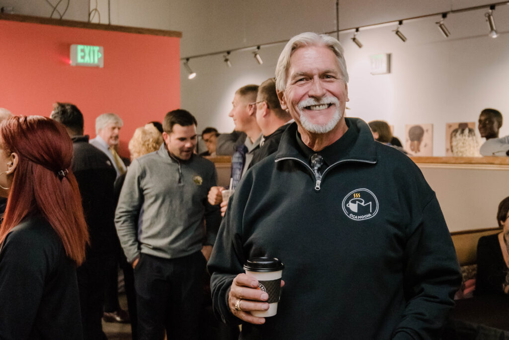 Man with white hair and a beard poses, smiling, holding coffee, wearing an OCA Mocha sweater, amidst a crowd.