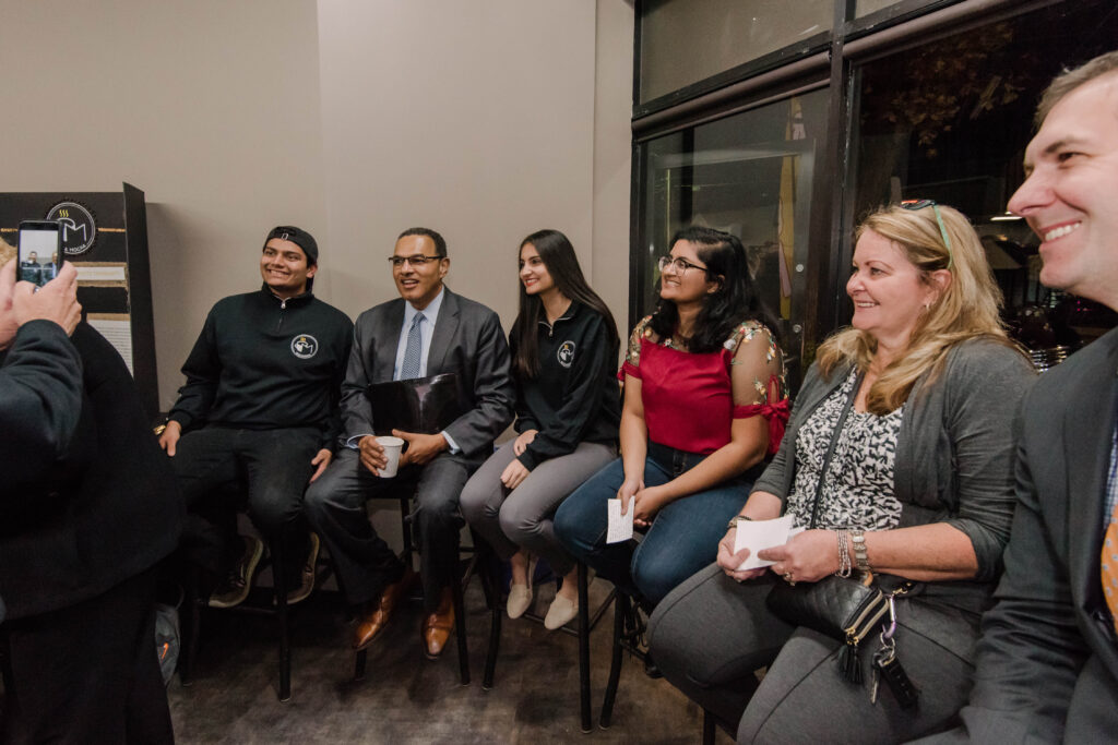 Six people of different ages sit together in a row, preparing to give remarks.