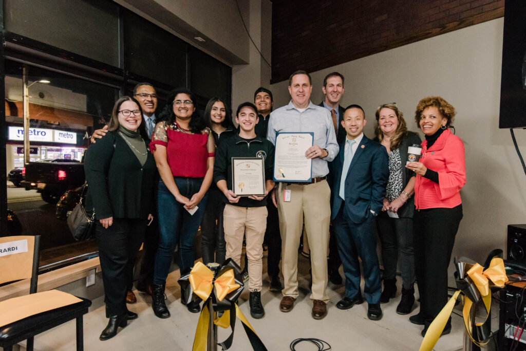 Eleven people pose on a small stage, holding local government citations in recognition of the space where they stand.