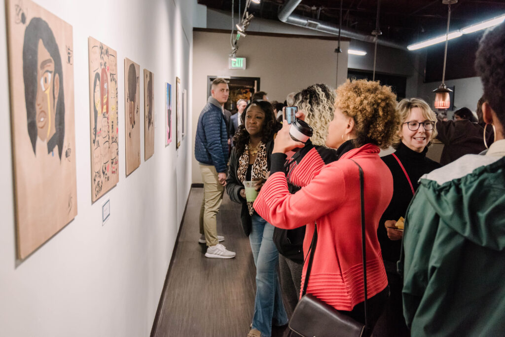 A woman takes a photo of a youth art exhibit in a busy community gallery space.