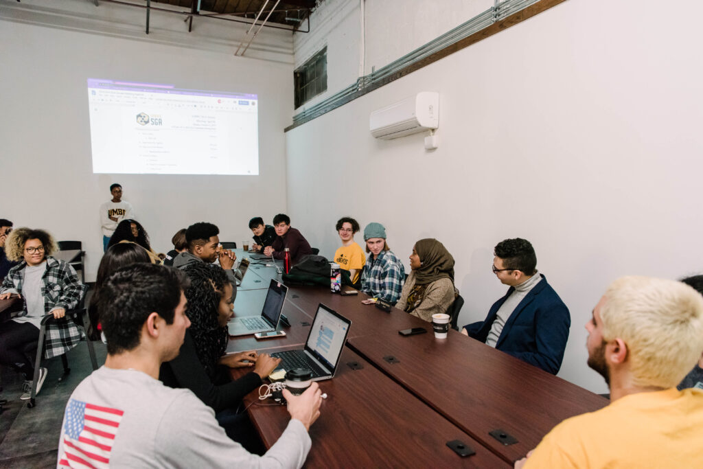 A group of young people gathers for a meeting around a table, with laptops.