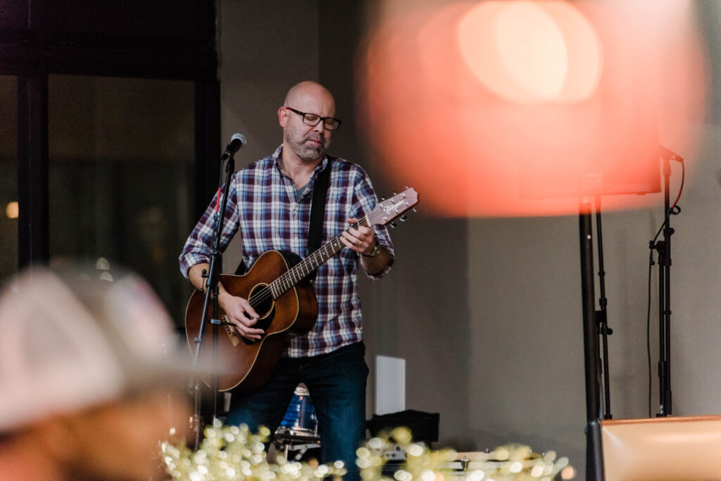 A man in a checked shirt and glasses plays a guitar onstage.