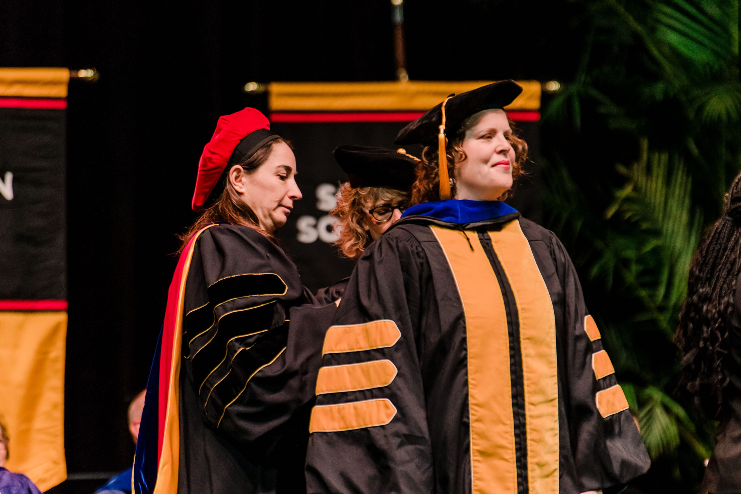 professors in graduation regalia help each other with outfit