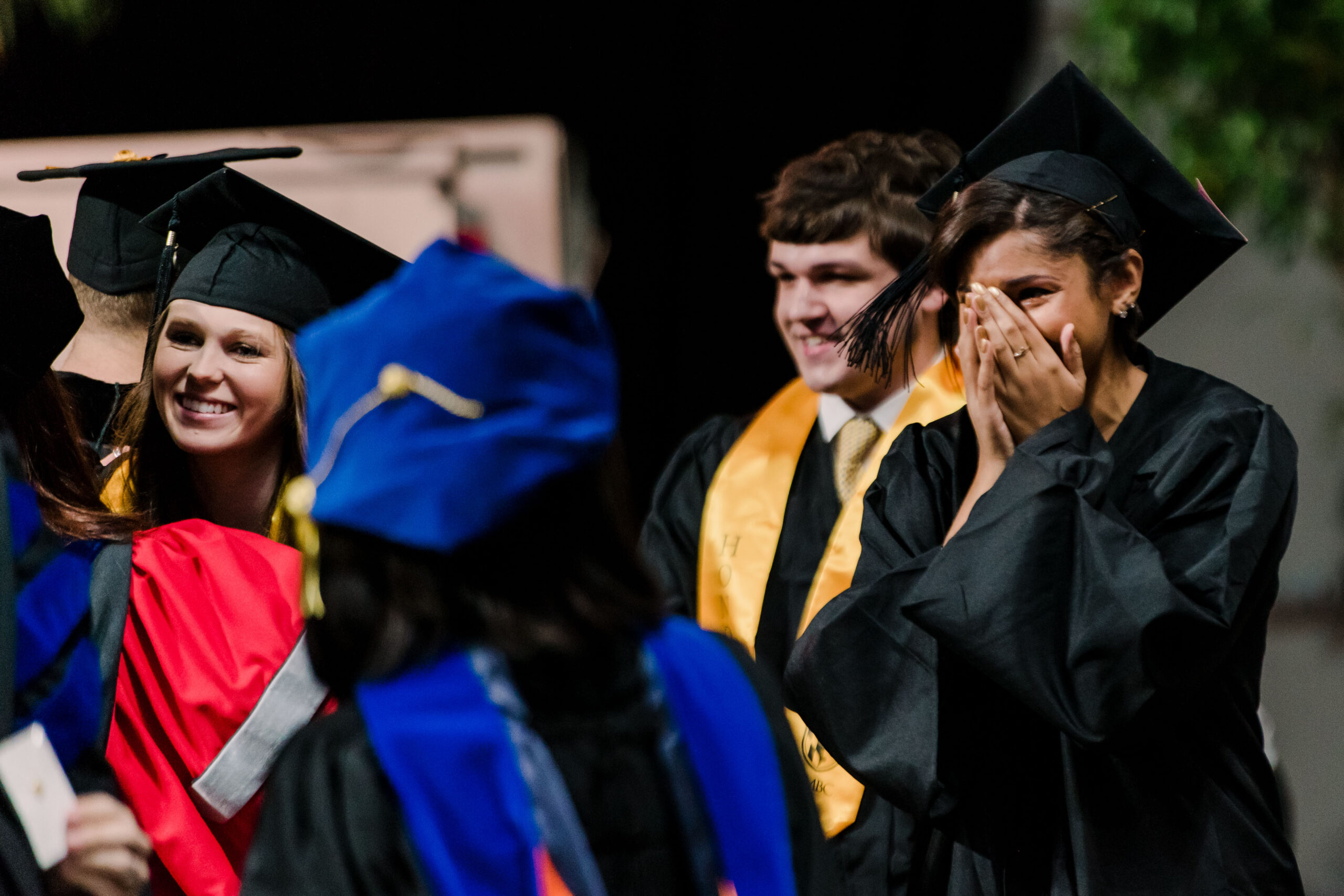girl surrounded by others in graduation garbs happily covers face