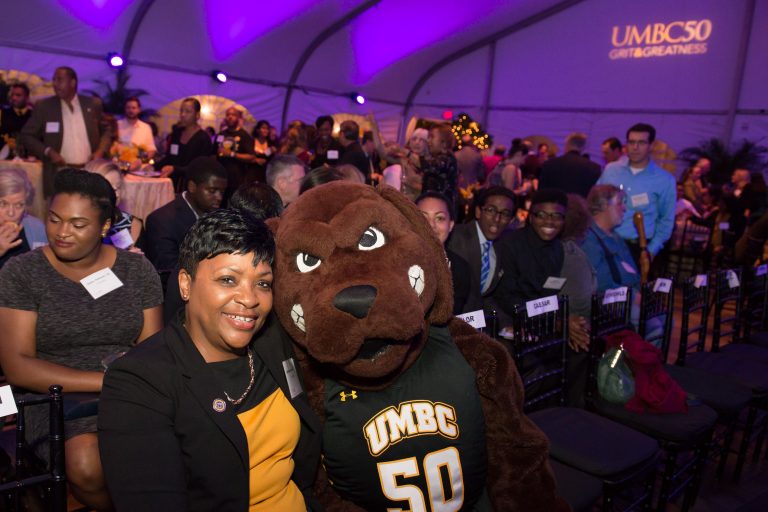 A black woman smiling with the UMBC retriever mascot at a celebration with many people in the background.