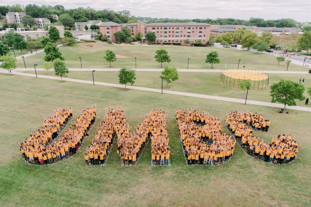 Hundreds of UMBC students gather in a field, standing in groups to spell out the letters "UMBC". They are wearing gold tops with black bottoms to match the school colors.
