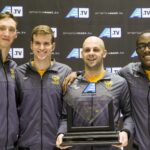 Four male swimmers wearing matching warm-up gear stand in front of an America East sign holding an America East trophy.