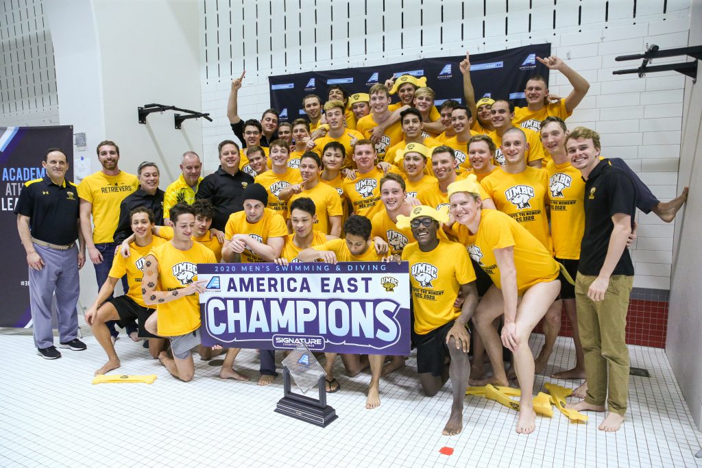 A men's swimming team gathers around a sign reading America East Champions