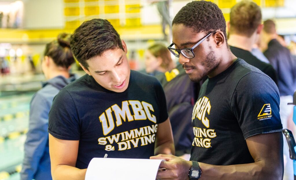 Two swimmers look at a paper together, wearing UMBC t-shirts next to a pool