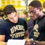 Two swimmers look at a paper together, wearing UMBC t-shirts next to a pool