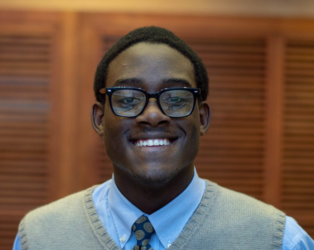 Student in glasses, a tie, and a sweater vest smiles in a portrait