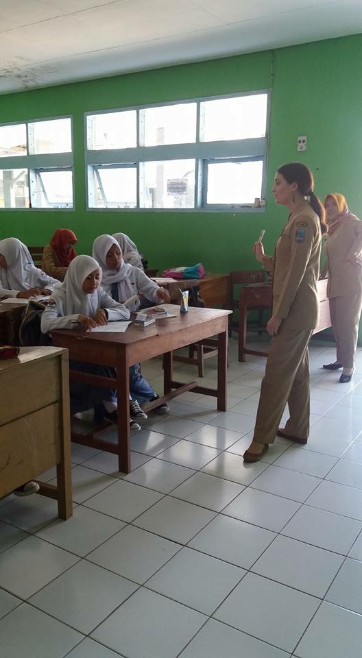 A young woman from the U.S. wearing a khaki uniform stands in front of a class of young teenage girls wearing white hijabs and sitting behind wooden desks in a bright green classroom with many windows along the side wall.